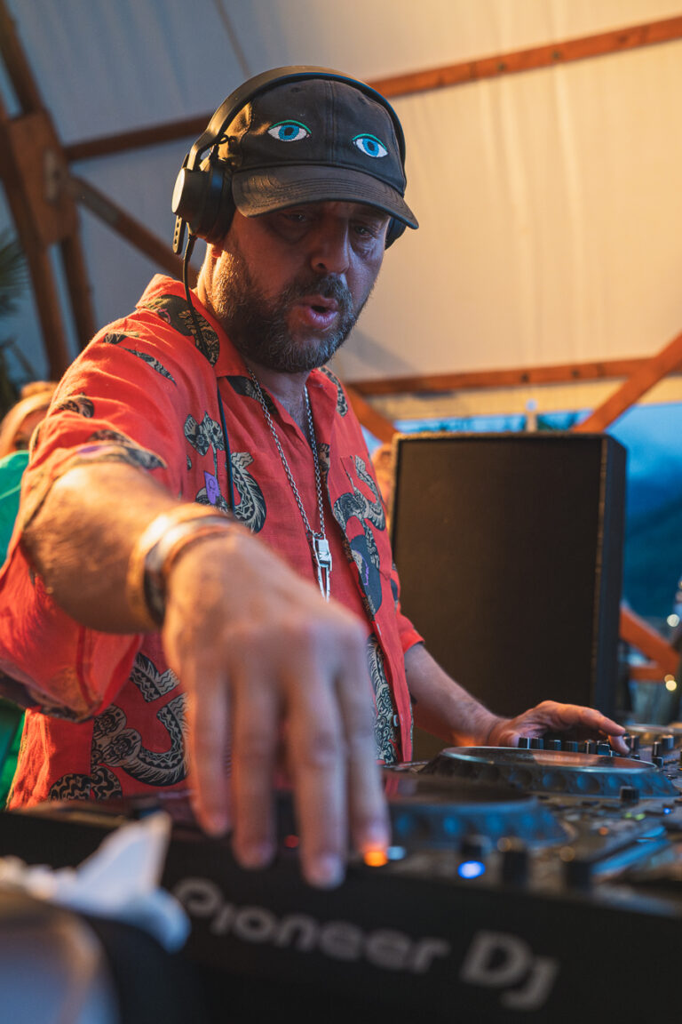 Ein bärtiger Mann mit Kopfhörern und einem gemusterten roten Kurzarmhemd steht hinter einem Pioneer-DJ-Mixer und bedient die Regler.