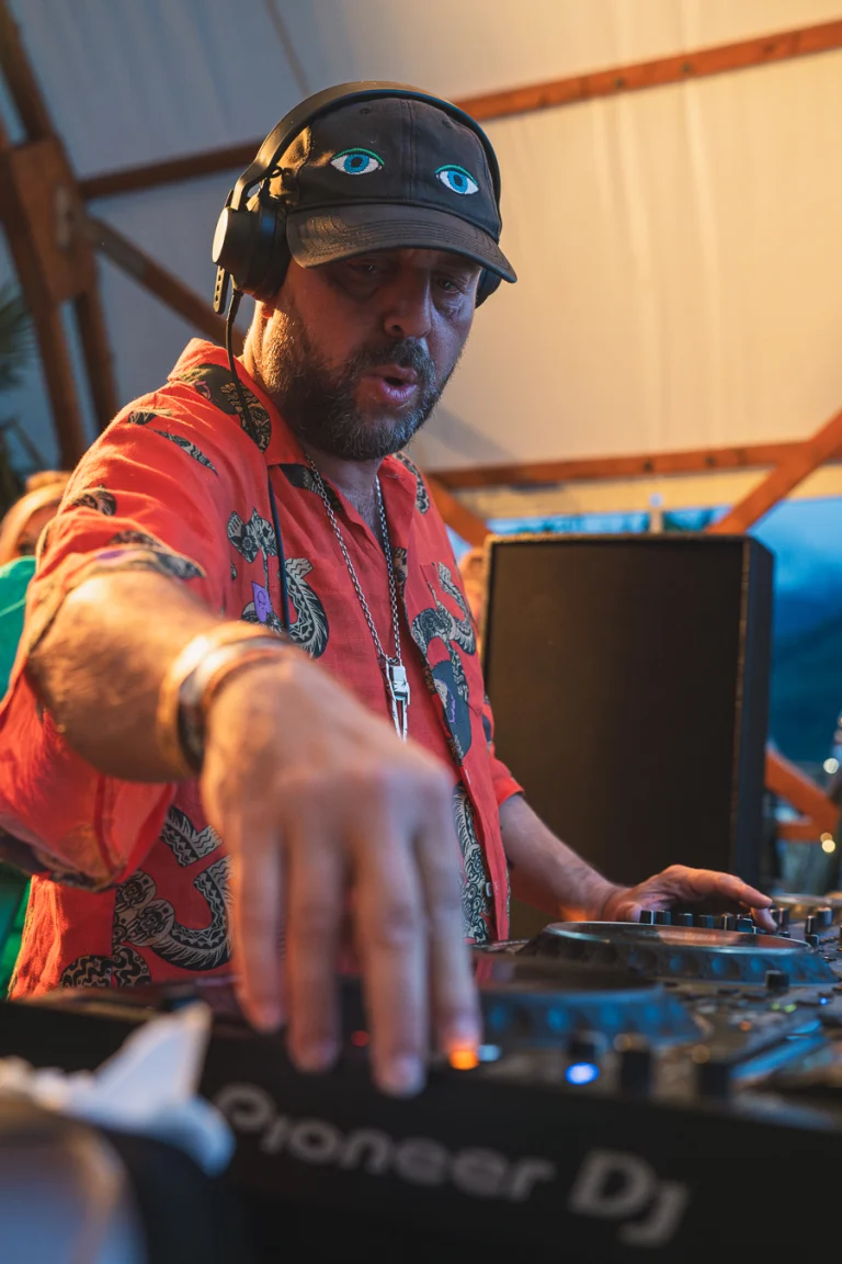 A bearded man wearing headphones and a patterned red short-sleeve shirt stands behind a Pioneer DJ mixer, operating the controls.