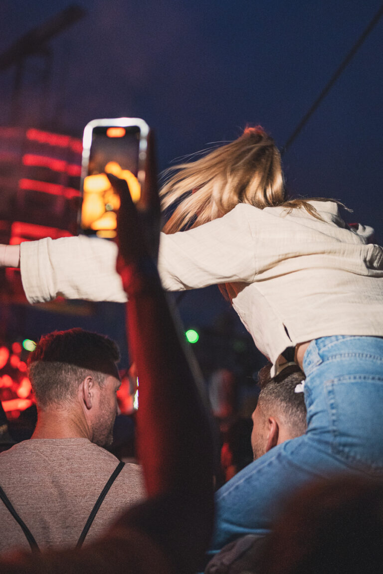 A woman sits on a man's shoulders in a crowd at night, raising her arms while another person in the foreground holds up a glowing smartphone.