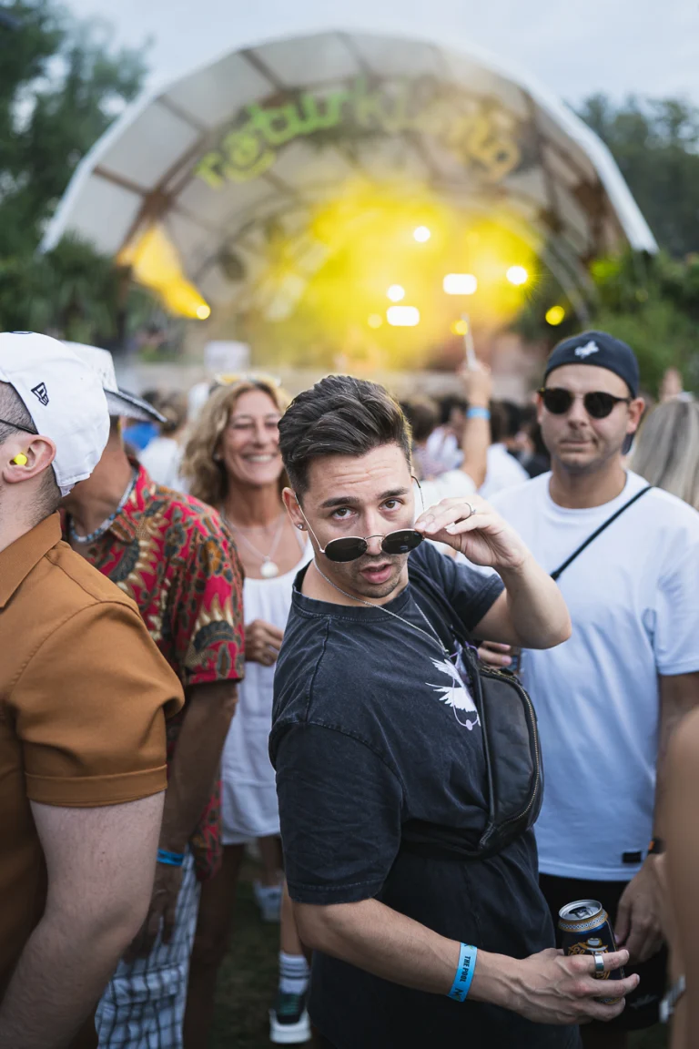 A man in the foreground adjusts his round sunglasses at an outdoor festival, with other attendees and a stage in the background.