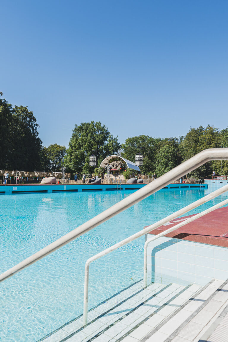 Ein Freibad unter klarem blauem Himmel mit Treppen und Metallhandläufen im Vordergrund sowie einer Bühnenkonstruktion im Hintergrund.