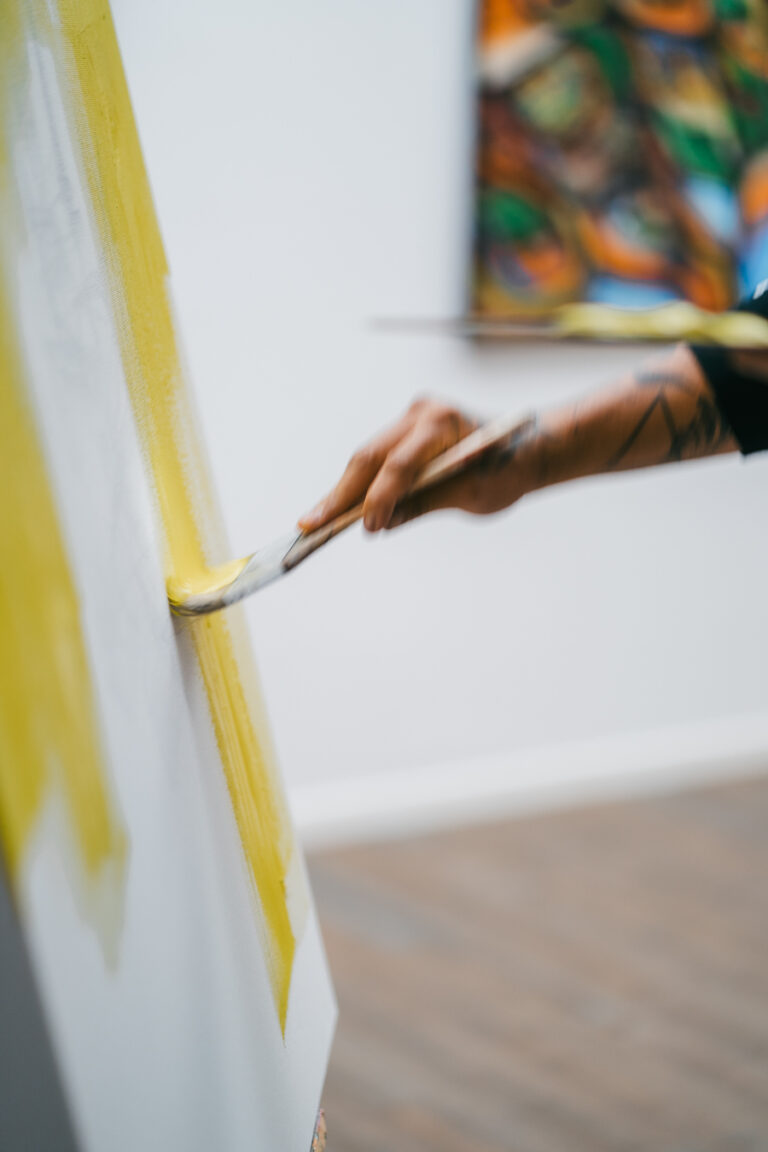 A person paints a yellow stroke onto a canvas on an easel.