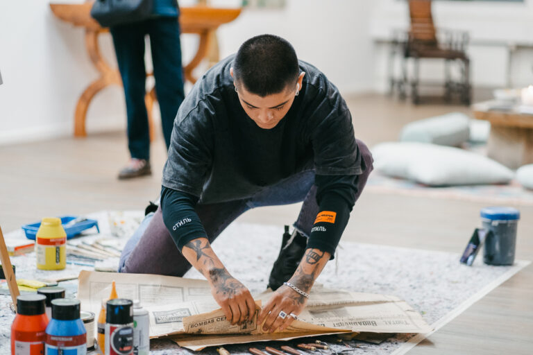 A tattooed man with short hair kneels on a studio floor, unrolling large sheets of printed paper amid paint bottles and tools.