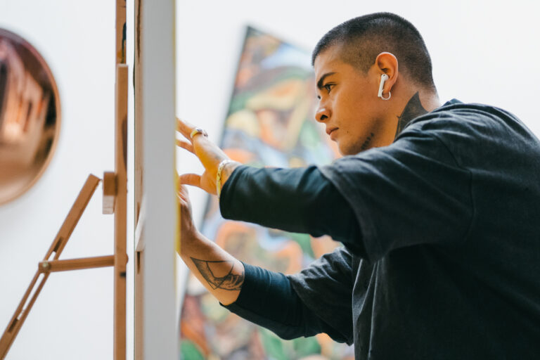 A tattooed man stands before an easel, carefully adjusting a canvas in bright indoor lighting.