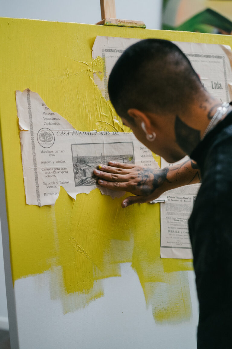 A man smooths a collaged newspaper fragment onto a yellow-painted canvas.