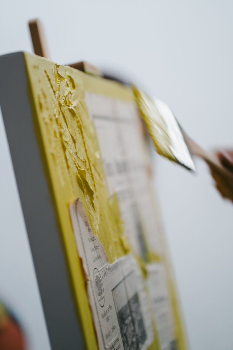 A hand applies thick yellow paint with a brush to a textured canvas collage.