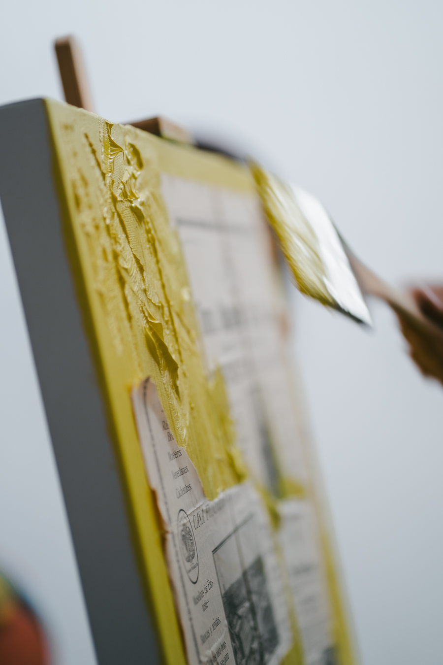 A hand applies thick yellow paint with a brush to a textured canvas collage.