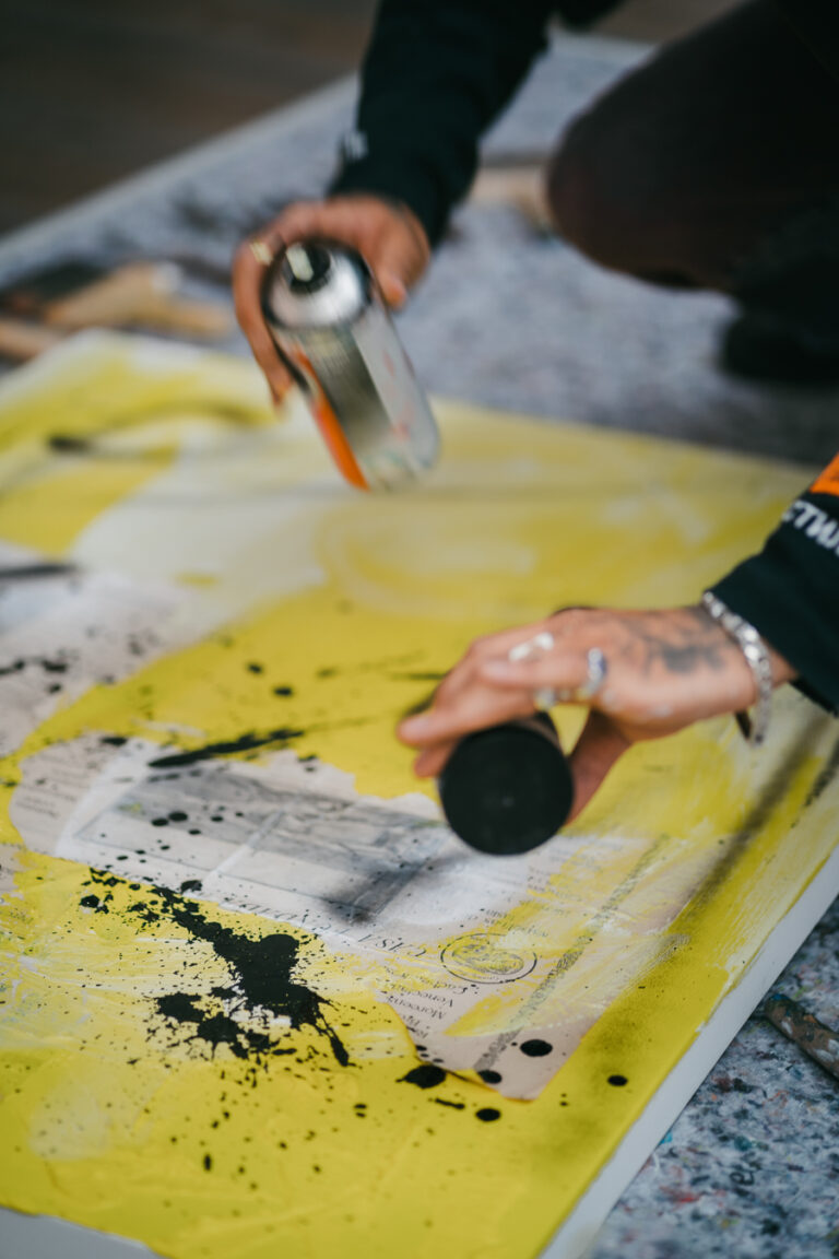A person kneeling over a large canvas on the floor, applying black spray paint from cans onto a yellow and printed surface.