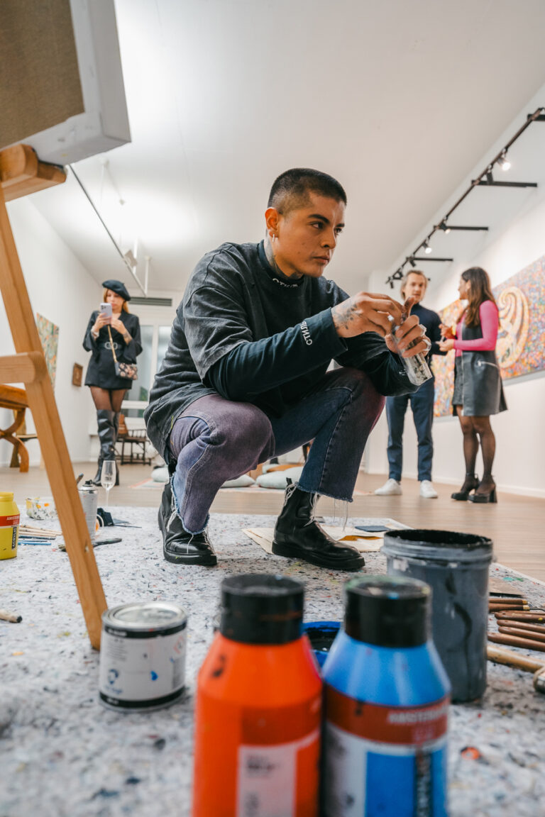 A man crouches in a bright gallery, holding a small brush, with paint bottles in the foreground and other people in the background.