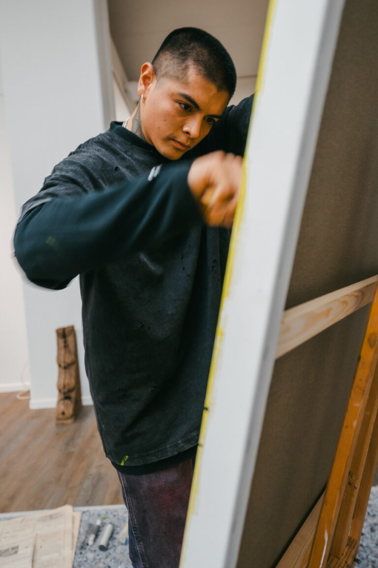 A man wearing a dark long-sleeve shirt works on a large canvas mounted on a wooden easel in an indoor studio.