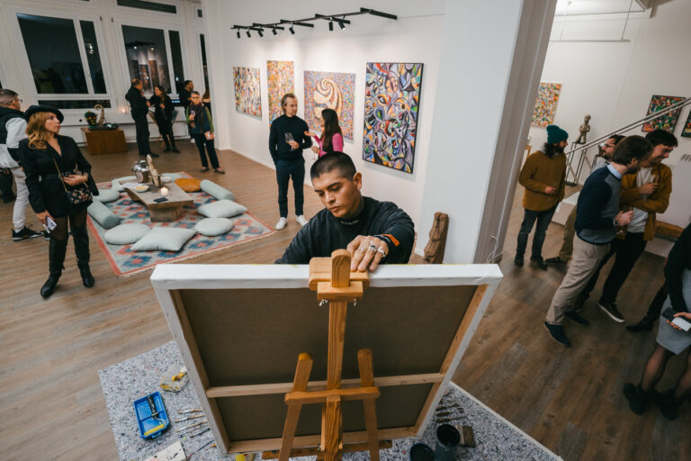 A man stands behind an easel painting a canvas in a brightly lit gallery space filled with visitors.