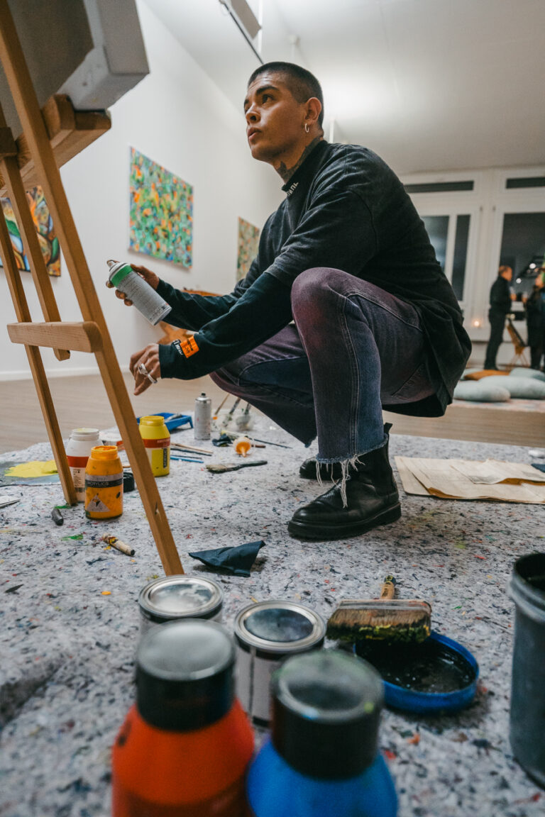 A man crouching in a studio, holding a spray can in front of a canvas on an easel, surrounded by paint cans and brushes on the floor.