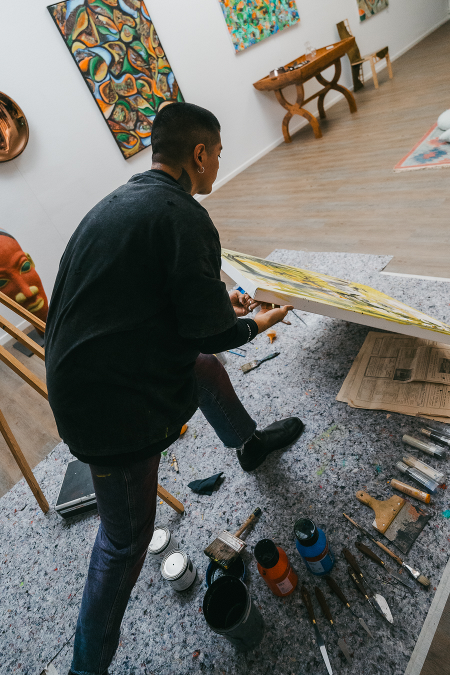 A man holds a large canvas in an art studio with paintbrushes, containers, and tools scattered on the floor.