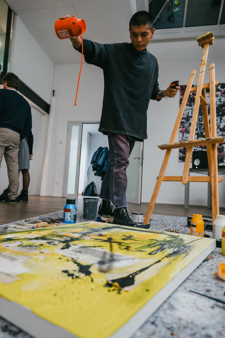 A man pours orange paint from a container above a canvas on the floor in a bright studio.