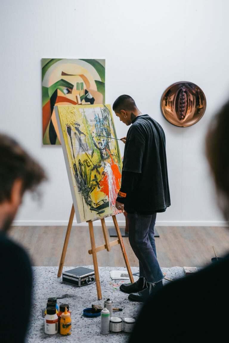 A man paints on a canvas on an easel in a brightly lit studio, while other people stand in the foreground.