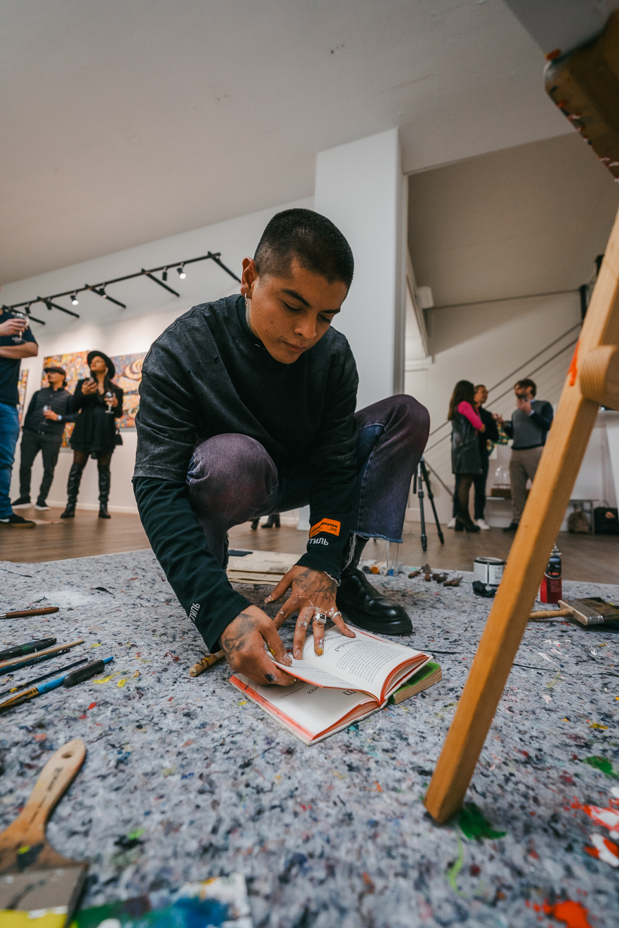 A man crouching on a paint-splattered floor inside a gallery, turning the pages of an open book.