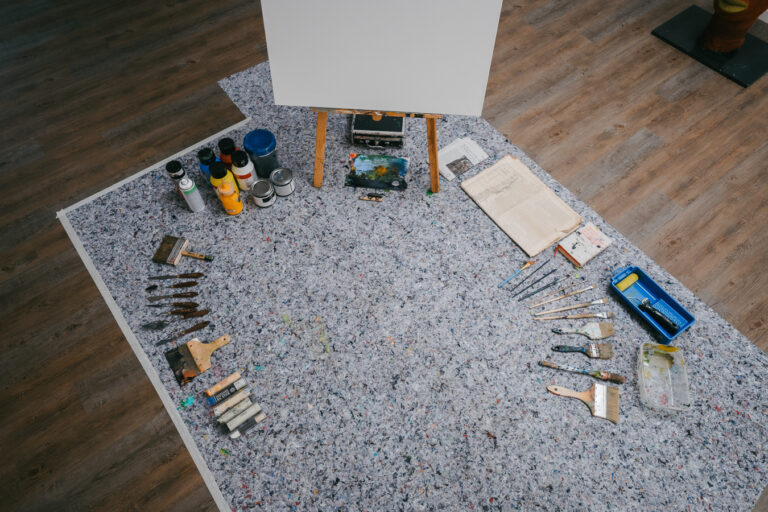 An empty easel stands above a drop cloth arranged with paint tubes, brushes, and palettes on a wooden floor.