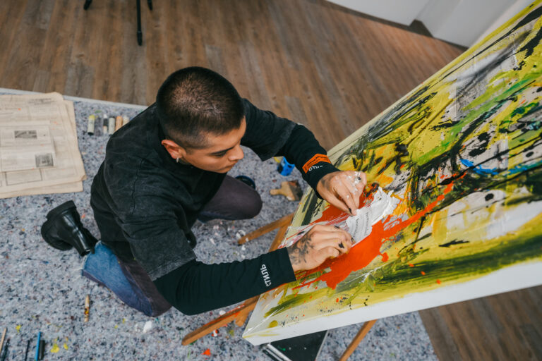 A man kneels on the floor, carefully applying white paint with his hands onto a large abstract canvas on an easel.