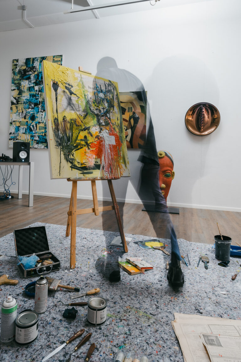 A motion-blurred figure of a person walks past an easel holding a brightly painted canvas in a studio.