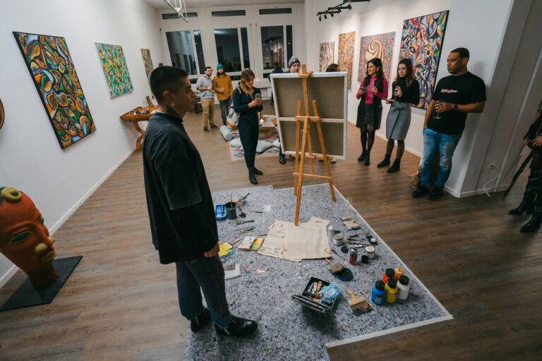 A man stands in an art gallery with paintings on the wall, looking toward an easel surrounded by art supplies while several people gather around.