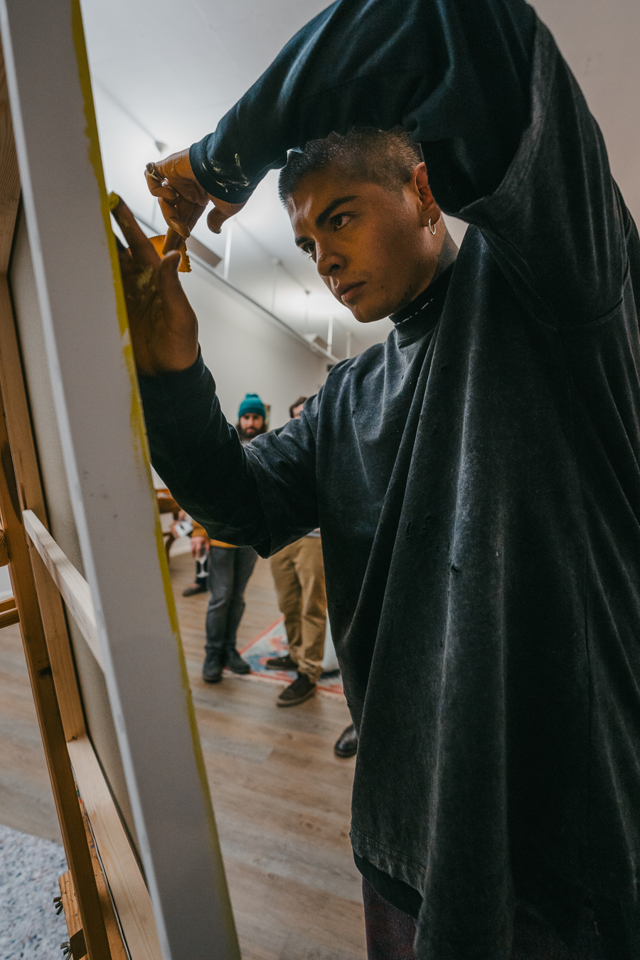 A man wearing a dark long-sleeve shirt paints on a canvas in an indoor setting while two other men stand in the background.