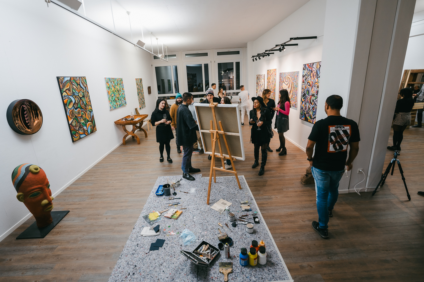 People gather in a bright art gallery with paintings on the walls and an easel with art supplies in the center.