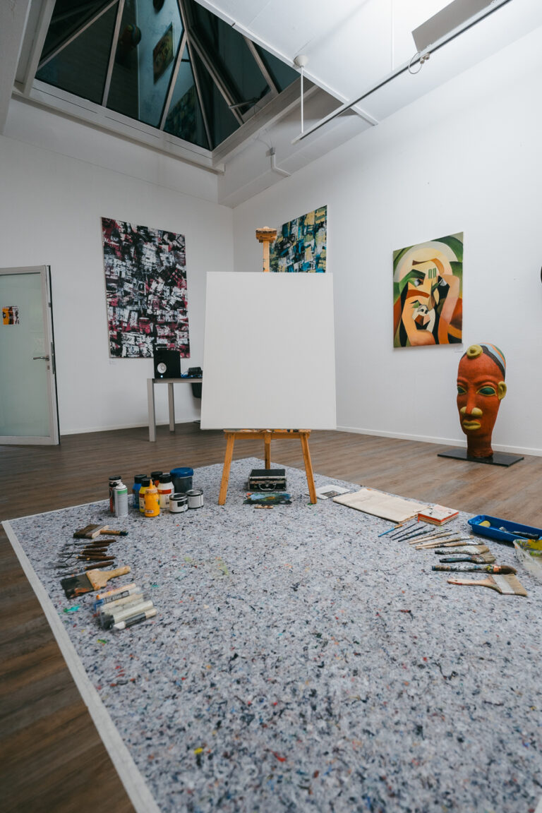 An empty canvas on an easel stands in an art studio, surrounded by painting tools laid out on a large grey rug.
