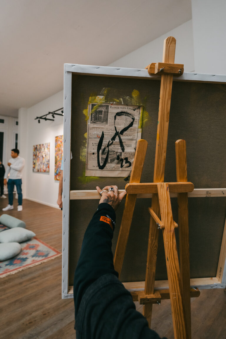 A person standing behind a wooden easel adjusts a canvas featuring an embedded newspaper clipping and abstract black brushstrokes in a gallery.