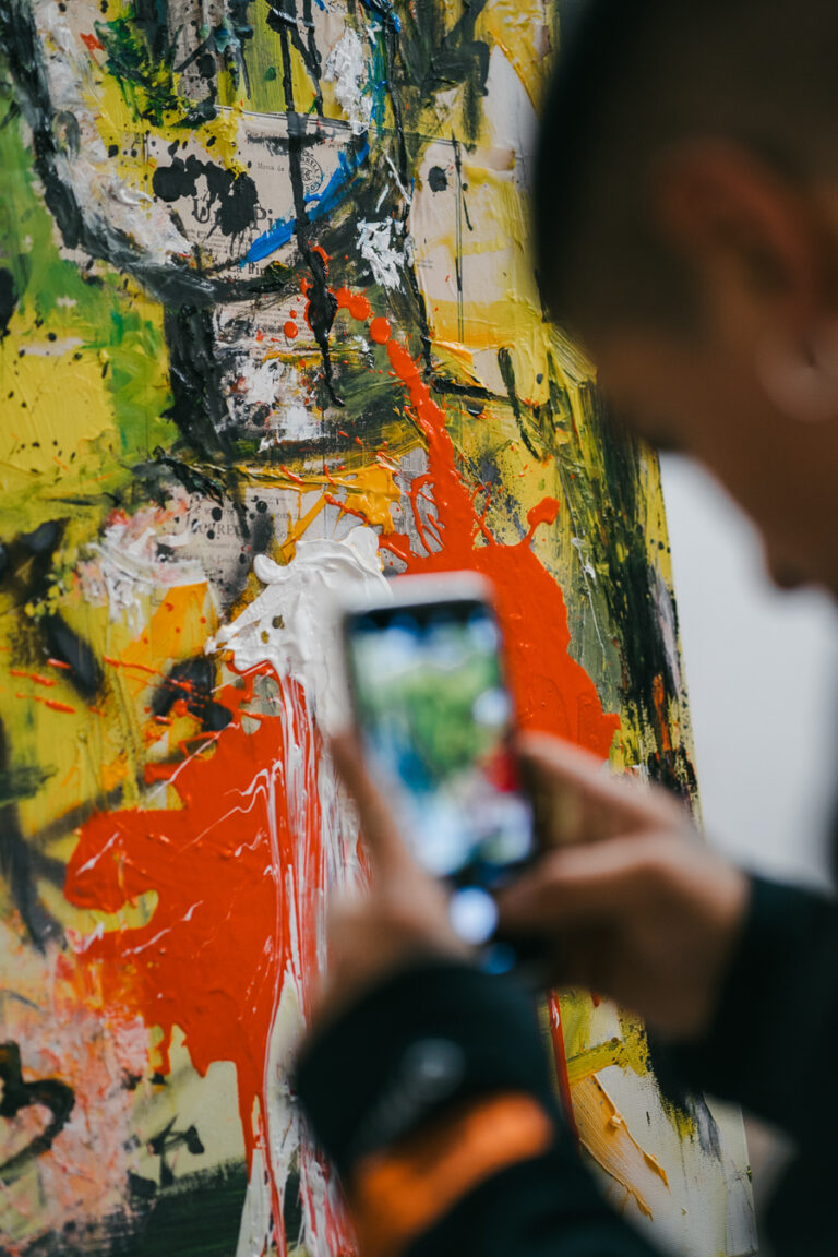 A man uses a smartphone to photograph an abstract painting with thick layers of yellow, orange, black, and white paint.