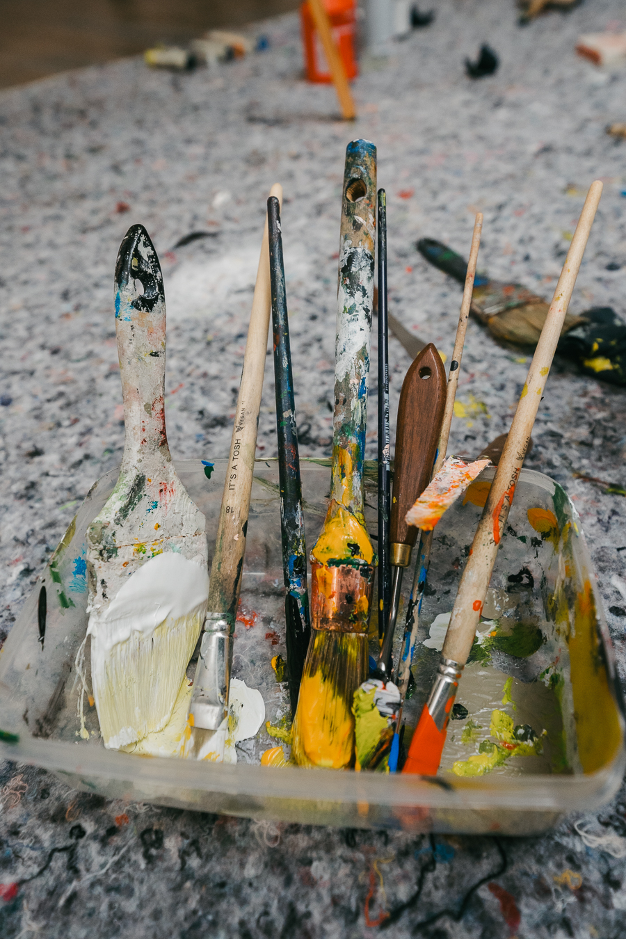 Various paint-splattered brushes stand upright in a clear plastic container resting on a speckled floor.
