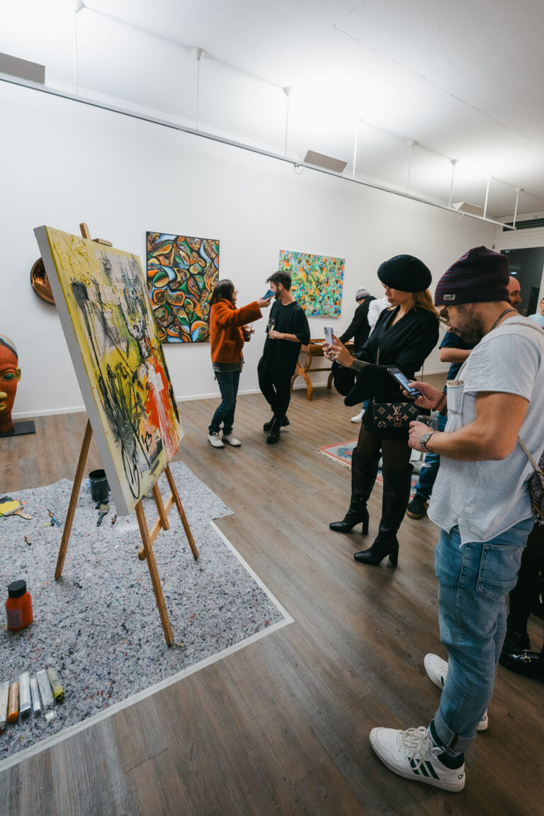 People view art pieces in a brightly lit gallery, including a painting on an easel in the foreground.
