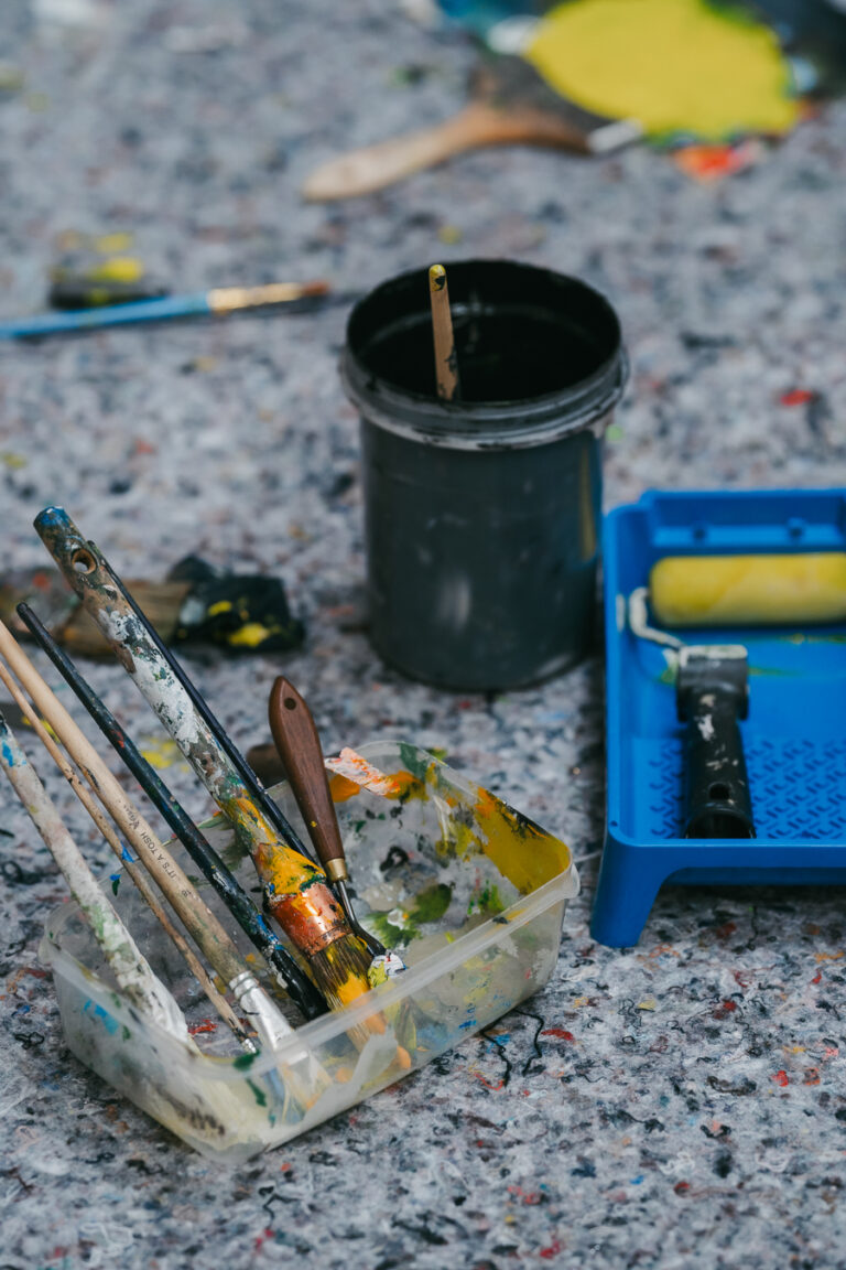 Various paintbrushes in a plastic container, a bucket, and a blue paint roller tray on a speckled floor.