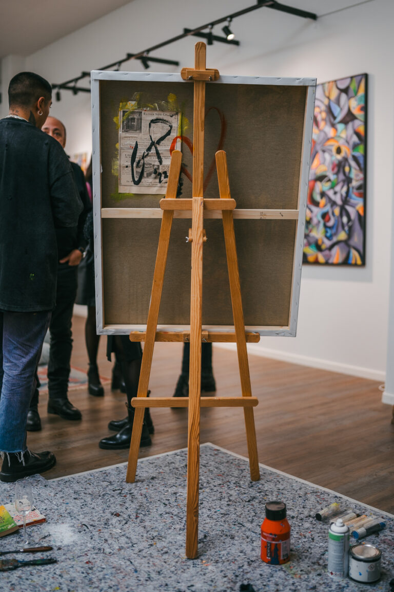 A wooden easel holding a canvas stands on a protective mat in an art gallery, with a man standing nearby and other people in the background.