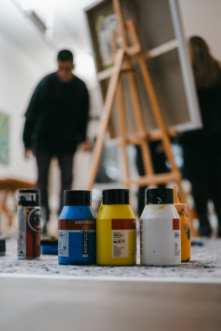 Paint bottles in blue, yellow, and white stand in the foreground while a man and a woman stand by an easel in a bright studio.