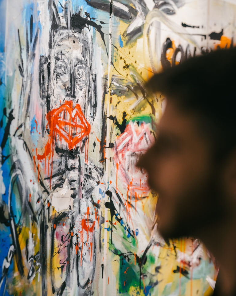 A blurred silhouette of a man in profile looks at a large expressive painting featuring abstract figures and bold splashes of red.