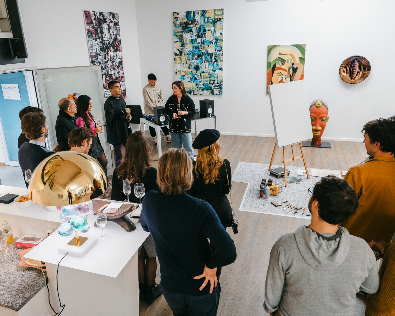 A woman speaks to a group of people gathered in a brightly lit art gallery, with paintings on the walls and a blank canvas on an easel.
