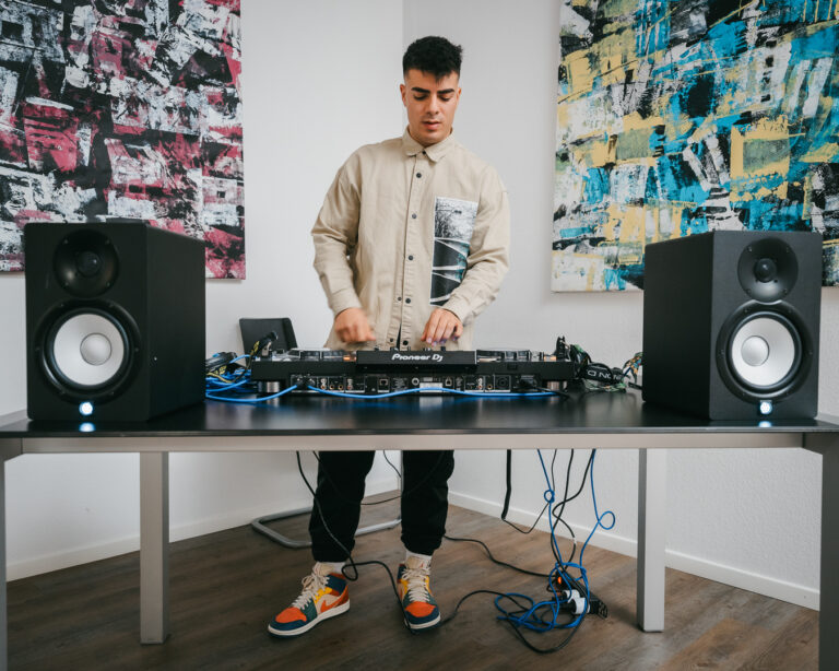 A man stands behind a desk using a Pioneer DJ controller, flanked by two large studio monitors, with abstract paintings on the white wall behind him.