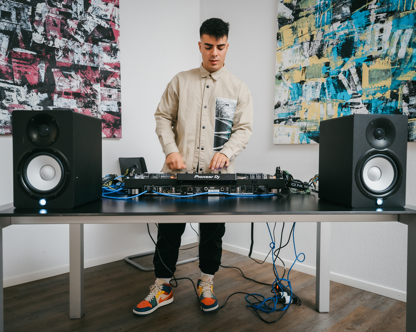 A man stands behind a desk using a Pioneer DJ controller, flanked by two large studio monitors, with abstract paintings on the white wall behind him.