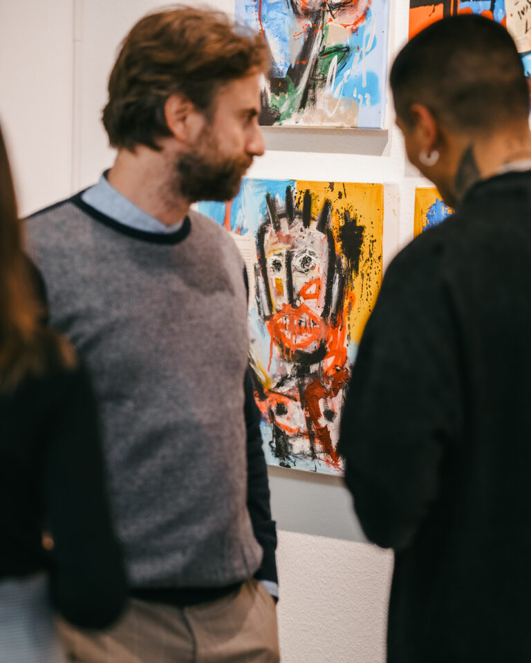 A bearded man looks at abstract paintings on a white wall while talking with another person.