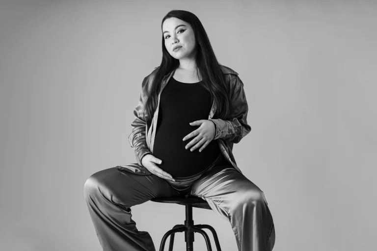 A pregnant woman with long dark hair sits on a stool, cradling her belly with both hands against a plain background in a black and white portrait.