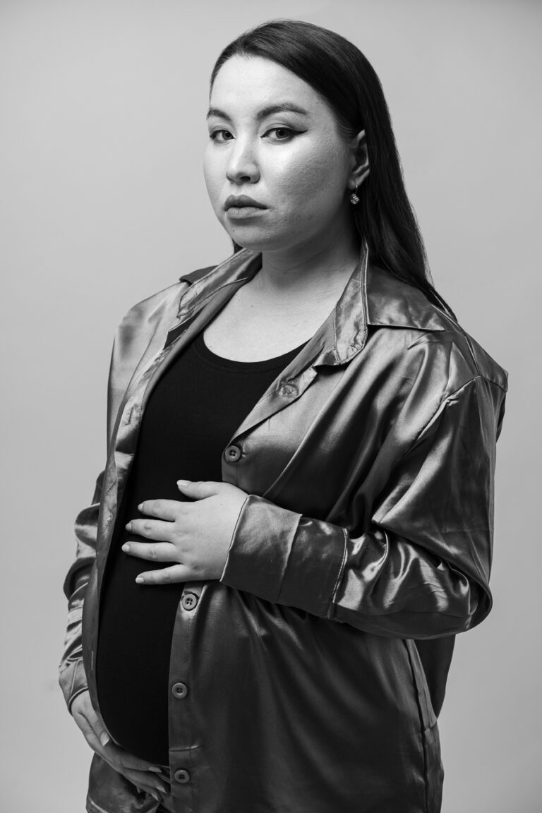 Black-and-white studio portrait of a pregnant woman wearing an unbuttoned satin shirt, holding her belly.
