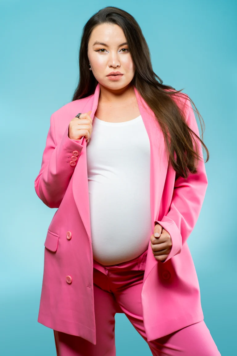 A pregnant woman in a pink blazer and white top poses against a bright blue background in studio lighting.