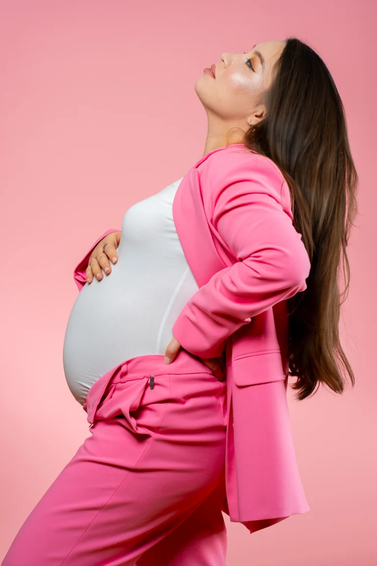 Profile shot of Guldana wearing a pink suit and white top, holding her pregnant belly against a pink background.