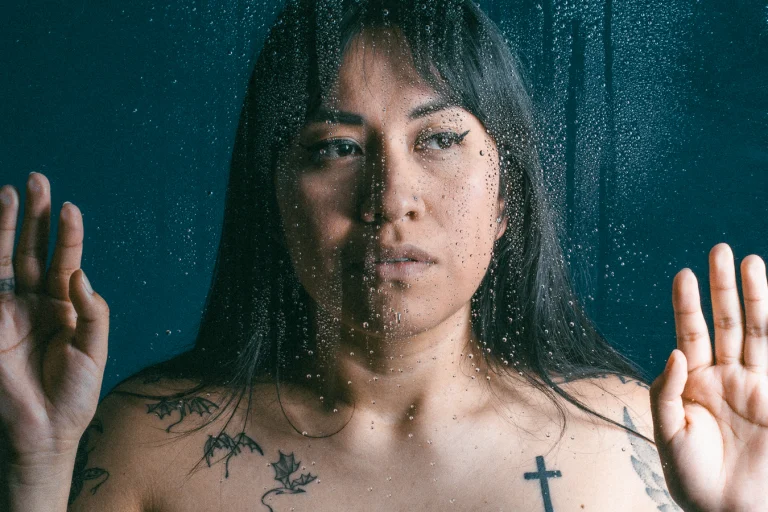 Portrait of Nina behind a glass surface covered in water droplets, with her hands pressed against the glass.