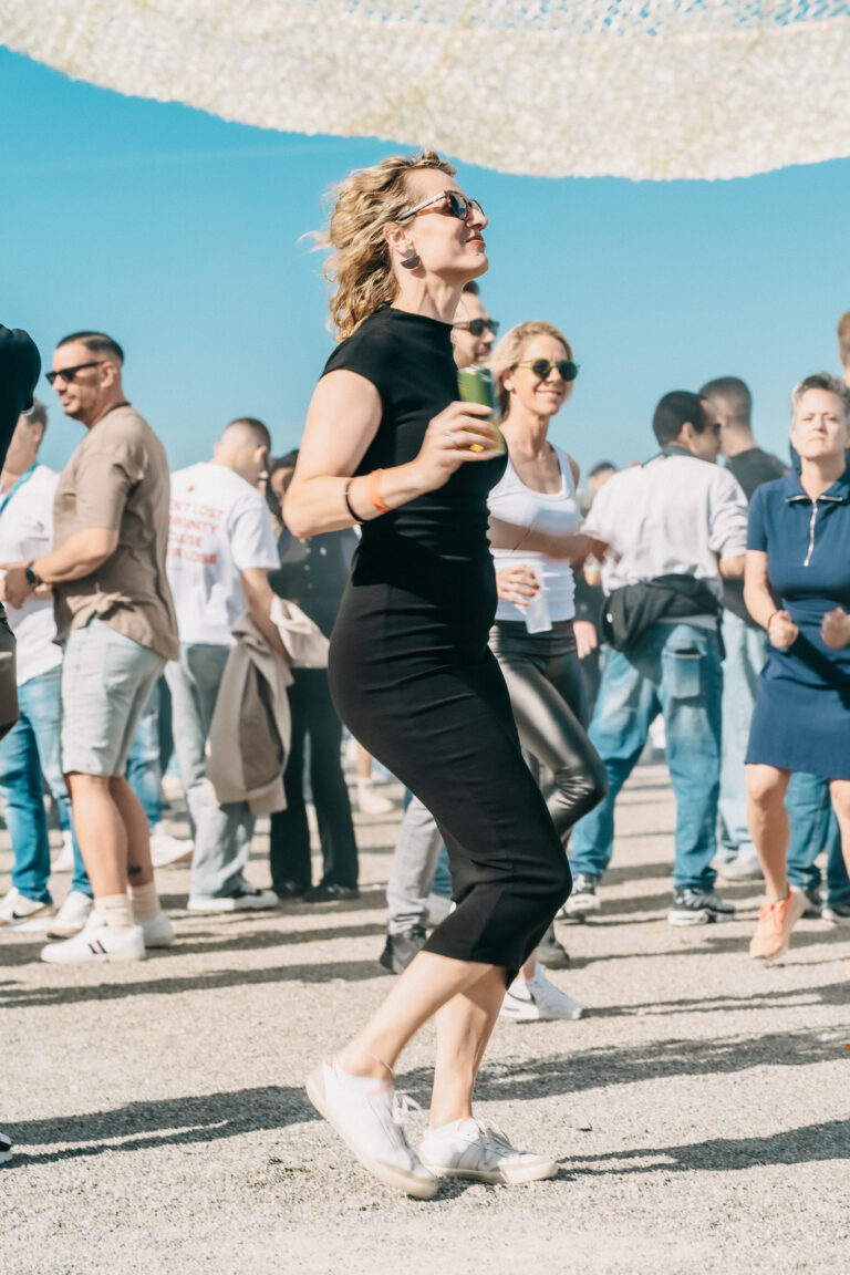 A woman in a black dress dances outdoors with a beverage in hand among a crowd under a bright sky.