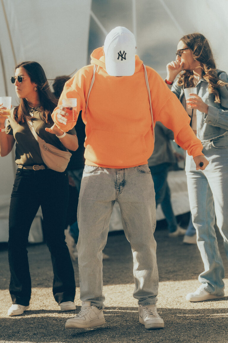 Ein Mann mit orangefarbenem Kapuzenpullover und weißer Kappe tanzt im Freien, flankiert von zwei Frauen mit Getränken.