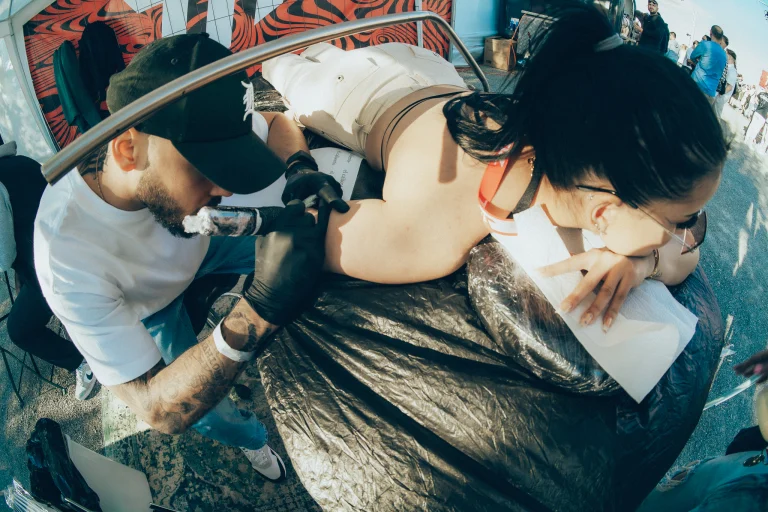 A man wearing a cap and black gloves tattoos a woman lying face down outdoors.