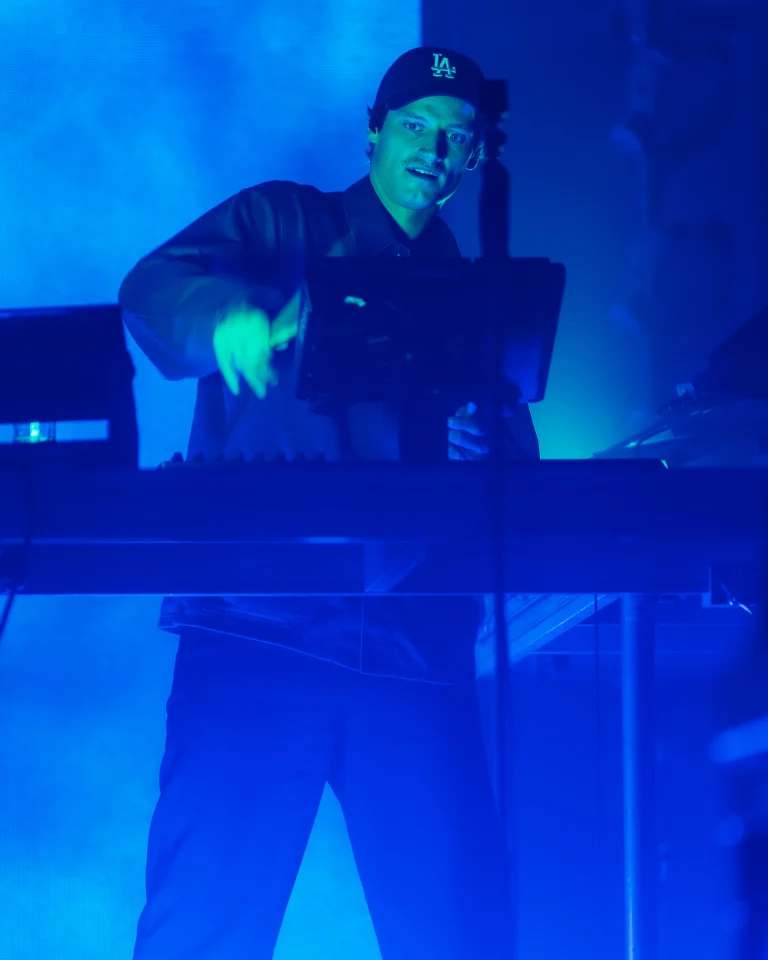 A man wearing a baseball cap stands behind a lighted mixing console, illuminated by blue stage light.
