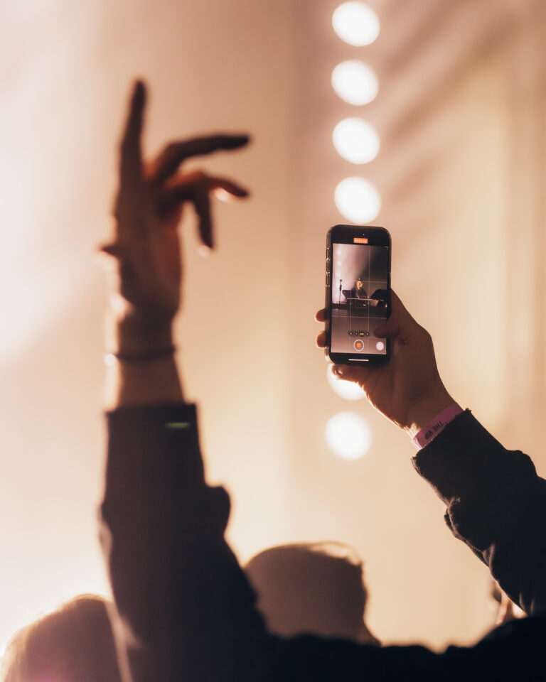 Eine Person hält ein Smartphone hoch, um ein Video aufzunehmen, während sie die andere Hand in einer Menschenmenge vor einem warm beleuchteten Hintergrund hebt.
