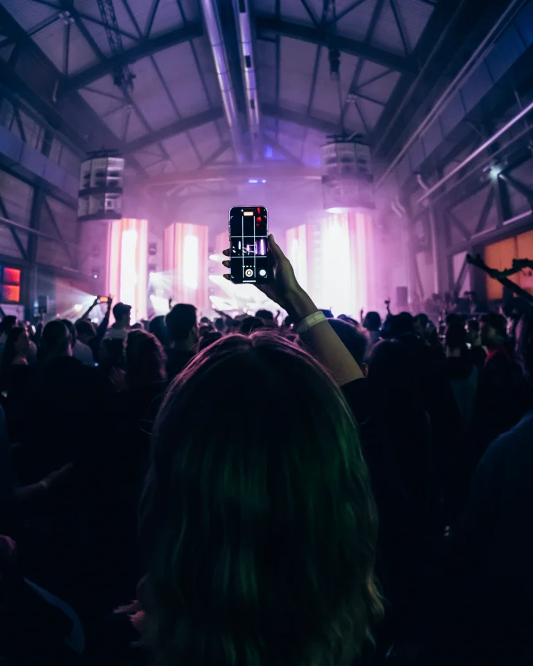 A woman holds up a smartphone to record a concert in a large industrial venue filled with a crowd under purple lighting.
